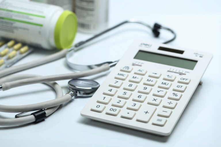 Calculadora, estetoscópio e medicamentos sobre superfície branca, representando a gestão de custos na saúde